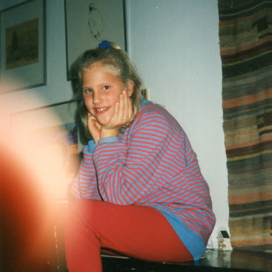 Toimittaja, kirjailija Venla Pystynen pikkutyttönä 1990-luvun alkupuolella kotona helsingin Pihlajamäessä. Istuu raitapaita päällään pöydällä ja katsoo kameraan.