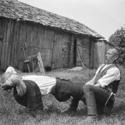 Två män leker leken "resa mast", där den ena ska försöka resa sig från liggande ställning i den andras famn utan att ta stöd av sina armar. Bilden är tagen 1929 i Strömfors, Kungsböle.
