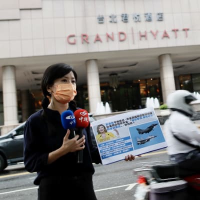 Taipein kadulla liikennevilinässä naisreporttertteri pitää mikrofonin lisäksi infokylttiä, jossa on hävittäjien kuvia ja nancy pelosi.