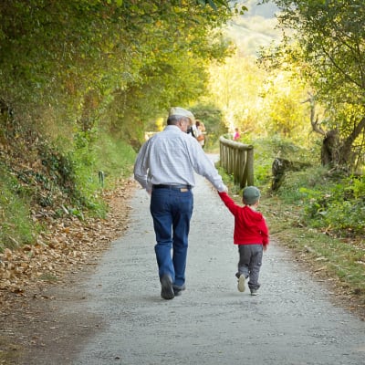 En farfar eller morfar håller sitt barnbarns hand medan de promenerar på en gångväg omringad av gröna träd och natur.