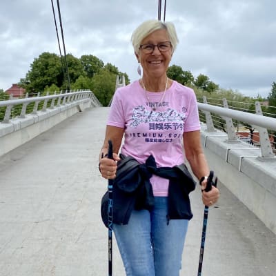 En medelålders kvinna i jeans och ljusröd t-skjorta stavgår över gångbron i Borgå. Hon tittar in i kameran och ler.