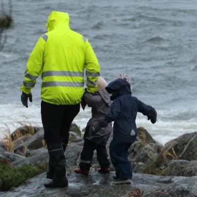 En pappa med två barn går på klippor i hård vind. Vattnet slår mot klipporna.