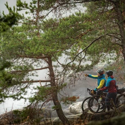 Två cyklister vid en strand med klippor och småväxta tallar. Den ena pekar ut mot havet. Båda bär cykelhjälm.