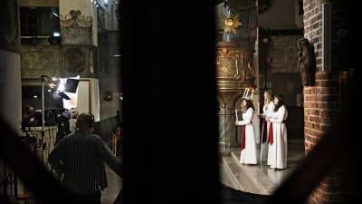 Bild tagen på långt håll. Luciatrio står framför ett altare, i bilden syns också män och tv-kamera.