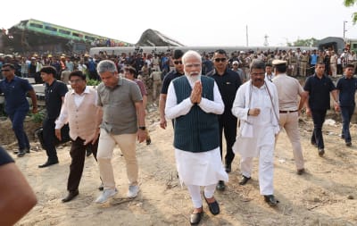 Narendra Modi hälsar med handflatorna ihoptryckta. Bakom Modi och hans entourage syns ett stort antal människor framför tågvagnar som sticker upp huller om buller.