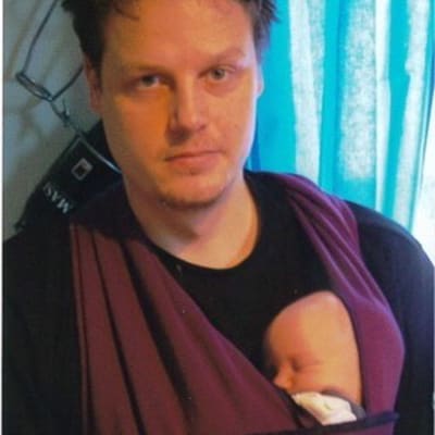 Teatterijohtaja Mikko Kanninen katsoo kameraan nukkuva esikoisvauvansa kantoliinassa rinnallaan.
