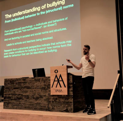 Fredrik Rusk föreläser på Åbo Akademi. Han står på ett podium och visar text på en stor skärm. Han har kort blont hår och skägg, och vit t-skjorta. 