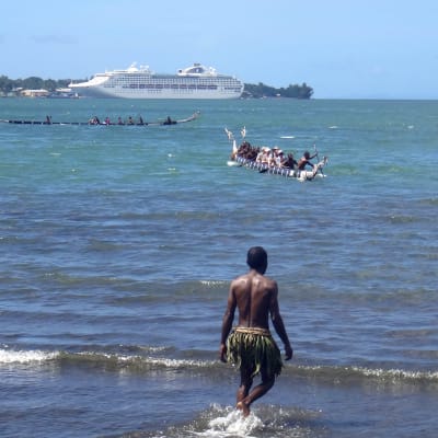 Personer i traditionella dräkter i Papua Nya Guinea. Bakom deras relativt primitiva båtar syns ett modernt kryssningsfartyg.