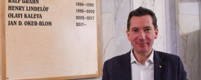En leende man i kostym bredvid en tavla på väggen. På tavlan står namnen på Lovisas stadsdirektörer, längst ner Jan D. Oker-Blom