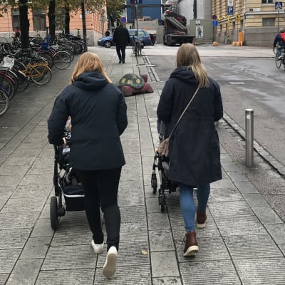 Två kvinnor promenerar med varsin barnvagn i stadsmiljö.