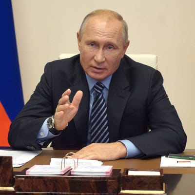 Rysslands president Vladimir Putin sitter vid ett sort skrivbord och pekar med hela handen. Den ryska federationens flagga står i bakgrunden.