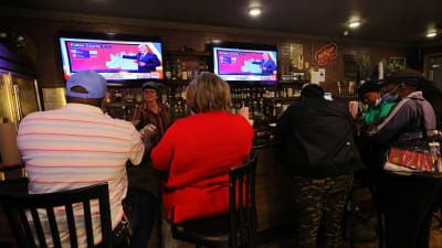 Människor sitter på barstolar vid en bardisk i Atlanta, USA. De tittar upp på en TV där man följer med rösträkning i senatsvalet i Georgia.