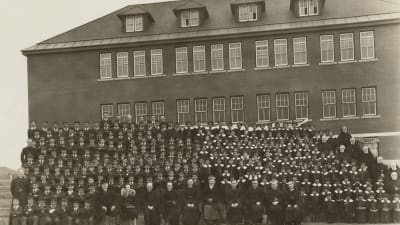 En svartvit bild av flera hundra elever på en läktare framför en stor skolbyggnad med en rad lärare i prästkläder på första raden.