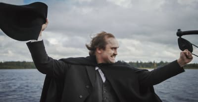 Man i kostym och svart mantel står och visar en segergest med en stor svart hatt i ena handen. I bakgrunden sjö och en strand kan skönjas längre bort.