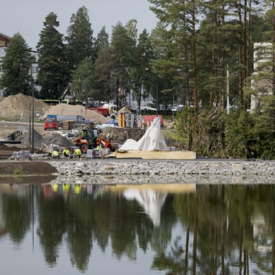 Matti Nykäsen muistomerkki nimeltä Höyhen odottaa palajastamista peitettynä Köyhälammen rannalla. Työntekijät rakentavat Köyhälammen puistoa. Köyhälammen pinta heijastaa peitetyn patsaan, ntylntekijät ja puita.