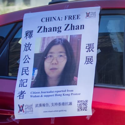 Två personer i munskydd sitter i en röd bil. På bilen hänger en lapp med en bild på journalisten Zhang Zhan och en uppmanan att fria henne från fängelset.