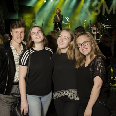 En kille och tre tjejer ler. De står framför en scen. Folk i bakgrunden.