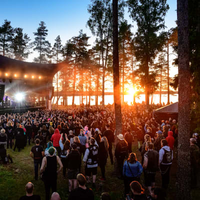 A crowd of people in a forested area by a lake, watching musicians perform on a stage at the Nummirock festival in 2021.
