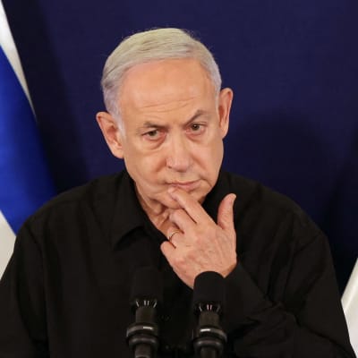 Benjamin Netanyahu i svart skjorta vid ett talarpodium med israeliska flaggor bakom sig. Han håller ena handen vid hakan och ser fundersam ut.
