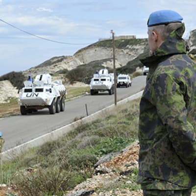 Vita pansarfordon och soldater på och intill en väg i ett rätt stenigt landskap. Soldaterna bär ljusblå baskrar och bilarna har texten UN.