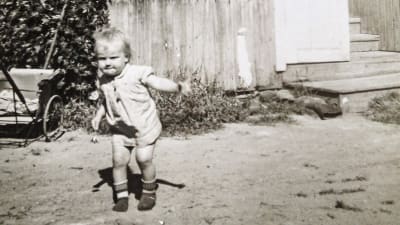 En svartvit bild på Stina som barn. Hon har en bestämd min och springer på gården framför en träbyggnad.