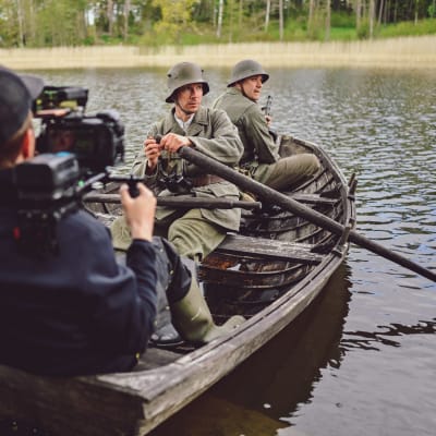 Två soldater i en gammal träeka. De är skådespelare. I båten sitter också en person i nutida kläder med filmkamera på axeln.