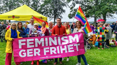 Personer står och ler med en banner där det står "Feminism ger frihet". I bakgrunden ett SFP-tält och prideflaggor.