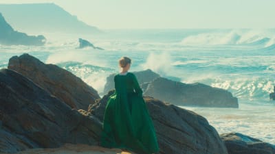 Nainen vihreässä puvussa seisoo selin kallioilla edessään myrskyävä meri, kuva elokuvasta Nuoren naisen muotokuva.