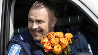 En polis sitter i en polisbil och tittar ut genom ett öppet fönster. Han ler och håller i en bukett gula rosor.