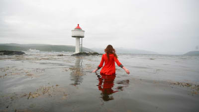 Kvinna i röd kappa vadar ut mot havet och fyren.