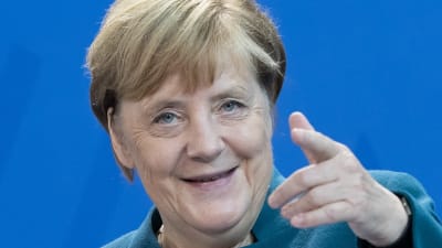 Angela Merkel pekar med pekfingret mot kameran och ler.
