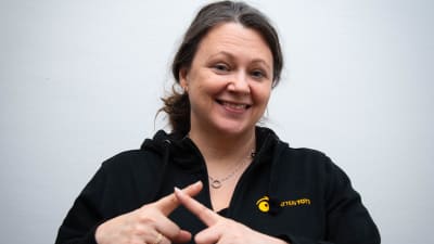Marita S. Barber håller båda pekfingrarna över varandra för teckent "stjärna". Hon ser rakt in i kameran och ler.