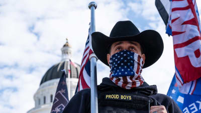 En man i cowboyhatt står framför Capitolium i USA. Han bär en scarf med USA:s flagga över ansiktet, på hans jacka står det "Proud boys".