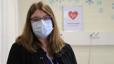 Kvinna med glasögon och munskydd tittar in i kameran. I bakgrunden sjukhusmiljö.