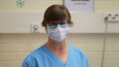 Kvinna i glasögon och munskydd och blåa vårdkläder tittar in i kameran.