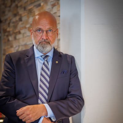 En medeålders, skallig man med grått helskägg och glasögon, i blå kostym. 