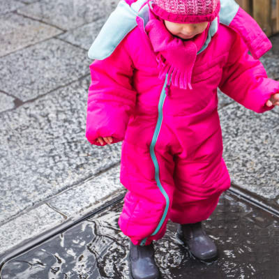 Ett barn i knallrosa vinterkläder och gummistövlar står i en vattenpöl