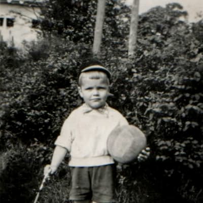 En lite pojke med en fotboll i ena och en promenadkäpp i den andra handen står framför en häck.