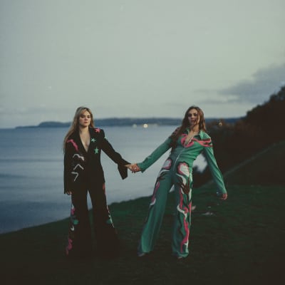 Två vuxna systrar håller varandra i hand på klipper med hav i bakgrunden.