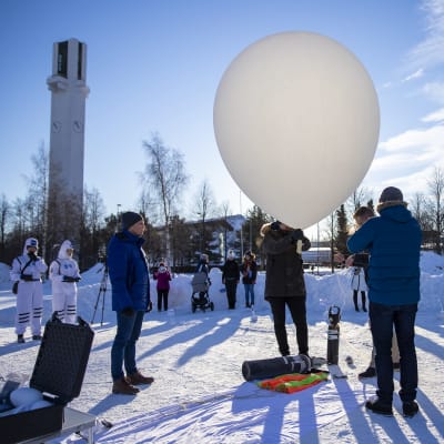 Joukko miehiä virittelee isoa valkoista ilmapalloa satelliitin kannattimeksi aurinkoisessa talvisäässä, runsaasti lunta ja Seinäjoen Lakeuden Risti näkyy taustalla.