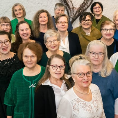 Joukko eri-ikäisiä naisia eli naiskuoro hymyilee kohti kameraa.