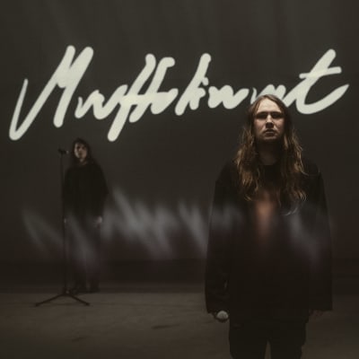 Kuvassa artisti Paperi T seisoo tummasävyisessä huoneessa. Taustalla lukee teksti "muuttolinnut".
