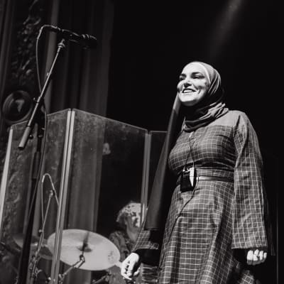 Sinead O'Connor katsoo lavalta yleisöön ja hymyilee. Hänellä on huntu päällään.
