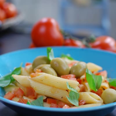 En portion penne pasta med tomatsås, garnerad med oliver och färska örter. Portionen är serverad på ett himmelsblått fat.
