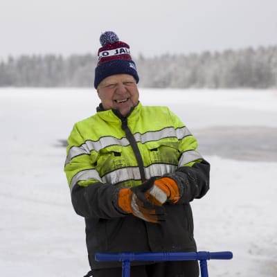 Naurava mies talvityövaatteissa lumisen järvimaiseman edustalla. 
