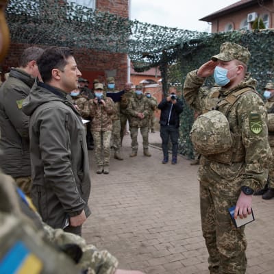 Ukrainas president Volodomyr Zelenskyj står tillsammans med en grupp soldater framför honom står en soldat i ansiktmask som gör honnör.