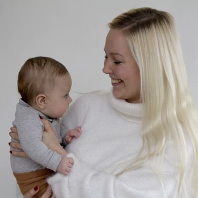 En kvinna med långt blont hår som håller en liten bebis i famnen. De tittar på varandra och hon ler. 