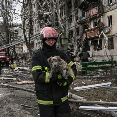 En brandman med en hund i famnen framför ett halvt raserat hus.