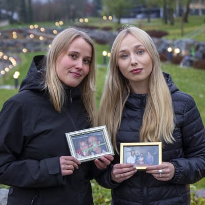 Två kvinnor (systrar) som står i håller i fotografier av deras föräldrar. 