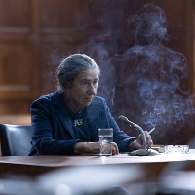 En rökande Golda Meir (spelad av Helen Mirren) sittande vid ett stort bord.